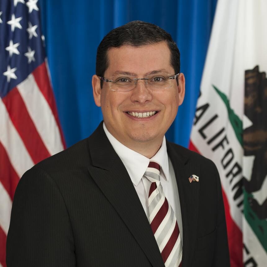 Headshot of Rudy Salas smiling in a black blazer and a striped red, white, and grey tie.