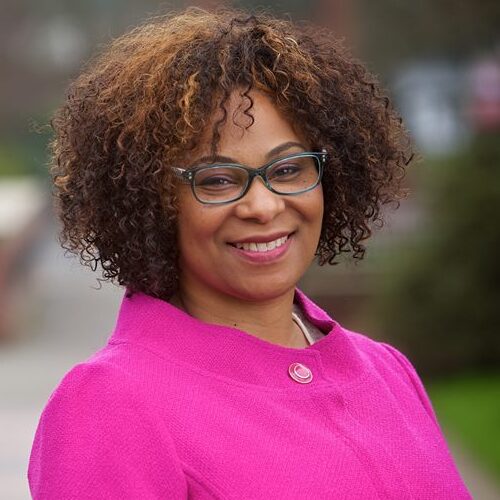 Headshot of Janelle Bynum in a pink top with long sleeves with voluminous curls, face framing glasses, and a smile.