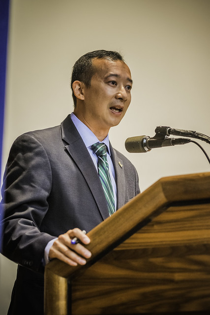 Lanon Baccam giving a speech in a grey suit, blue shirt and green striped tie while casually holding a pen in his right hand.