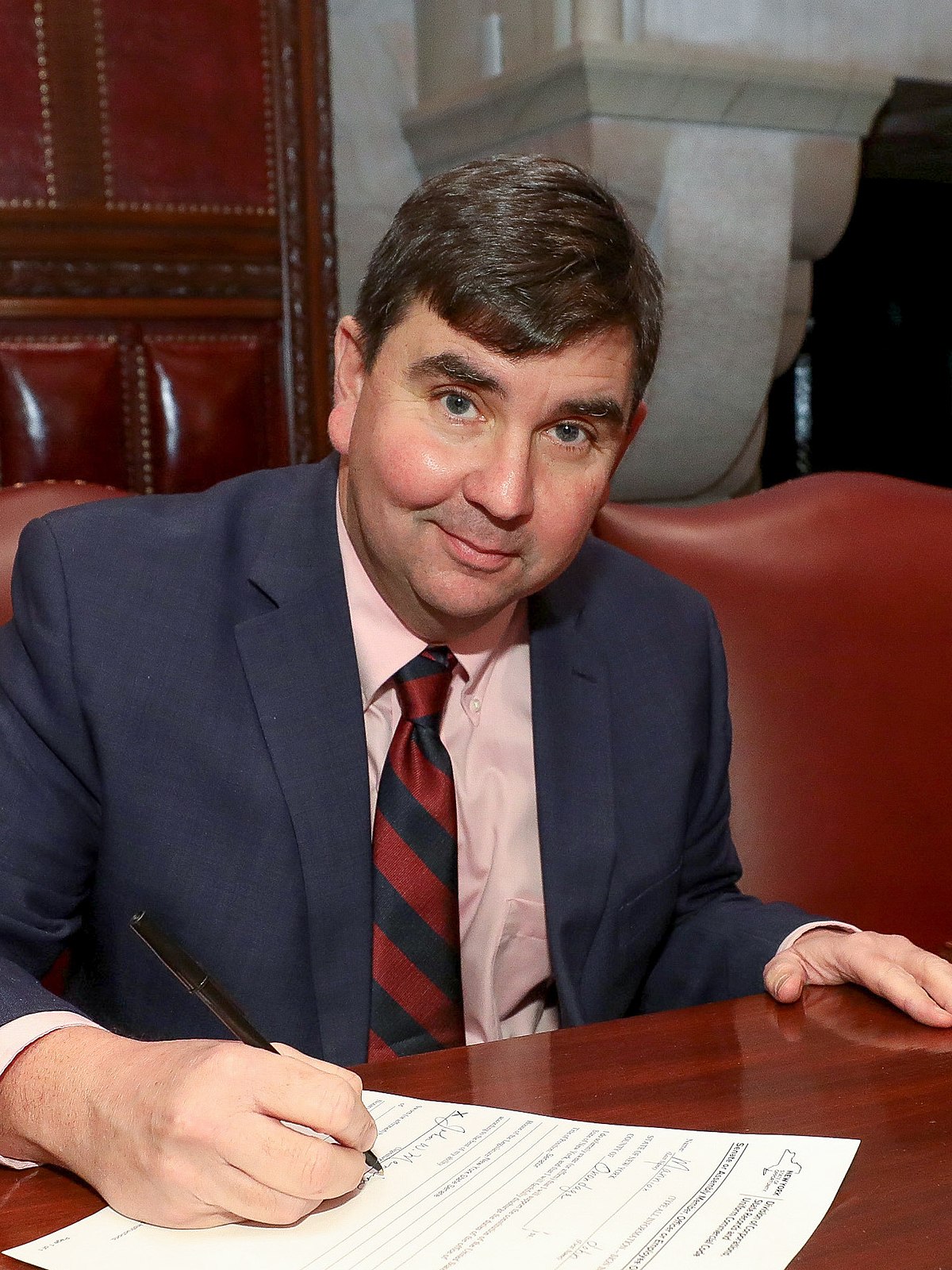 John Mannion in a blue blazer smiling at the camera while signing a document.