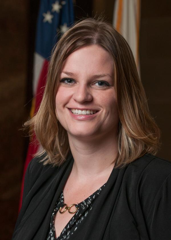 Headshot of Katrina Shankland in all black with a flag placed in the background.