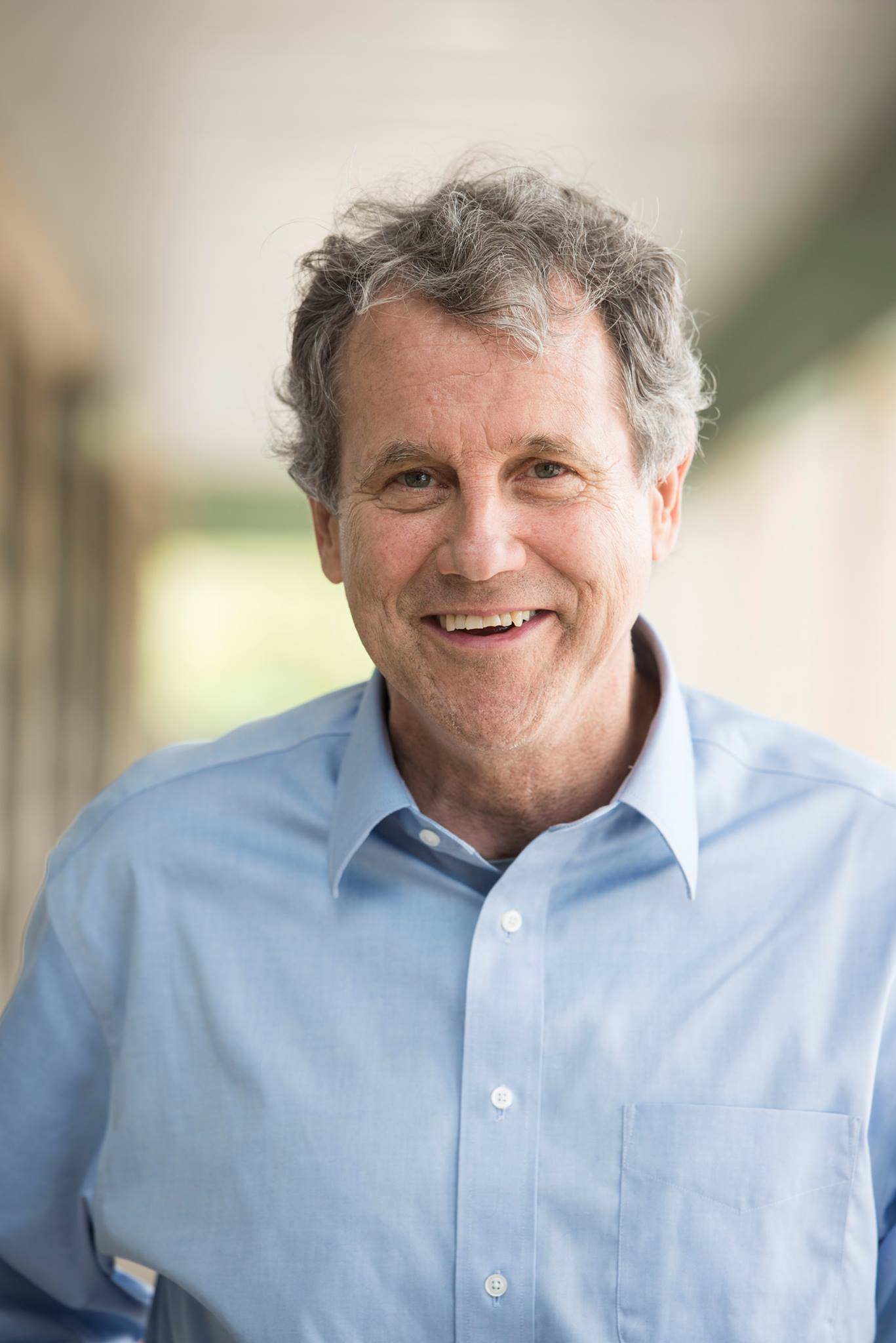 The image shows Sherrod Brown smiling warmly while wearing a light blue button-down shirt. His hair is slightly tousled, and the background is softly blurred, giving the image a relaxed and approachable feel.