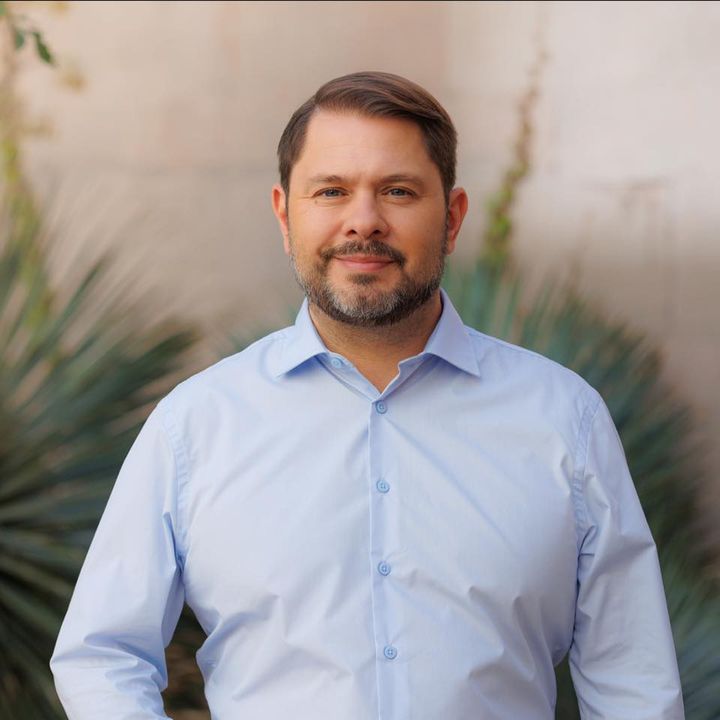 Ruben Gallego stands confidently in a light blue shirt with a desert landscape in the background.