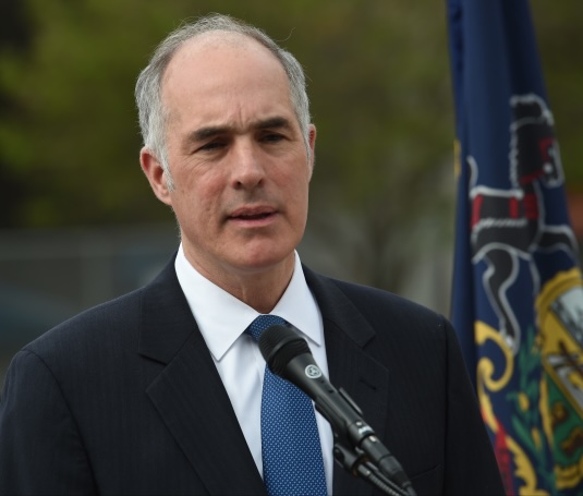 Bob Casey Jr. in a navy blue suit with a light blue tie. He's infront of a microphone in the middle of a speech.