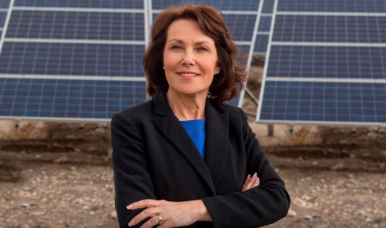 The image shows Jacky Rosen standing confidently with arms crossed in front of a backdrop of solar panels. She is wearing a black blazer over a blue top, and her expression is warm and determined.