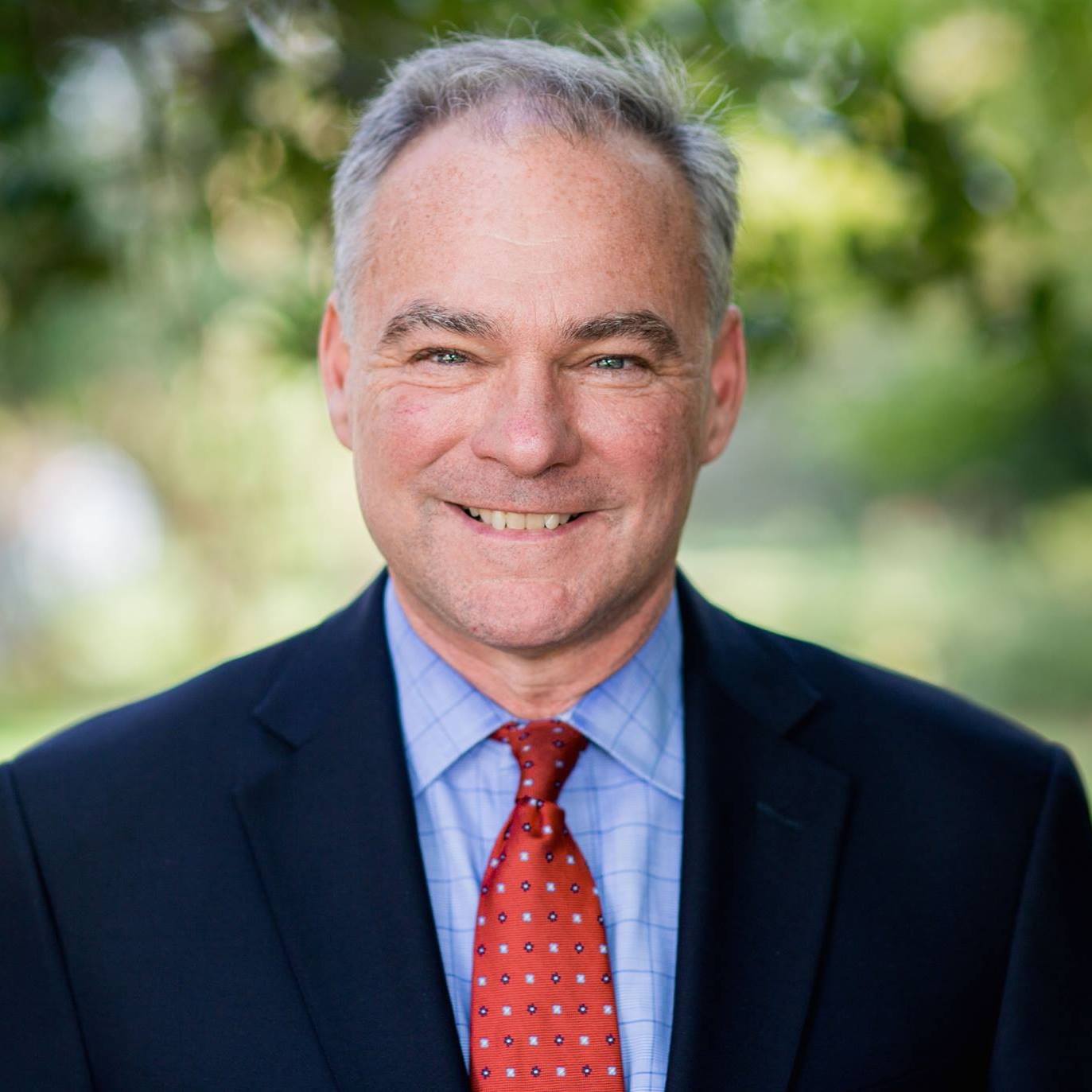 Tim Kaine in blazer with a red tie on in front of a blurred greenery background.