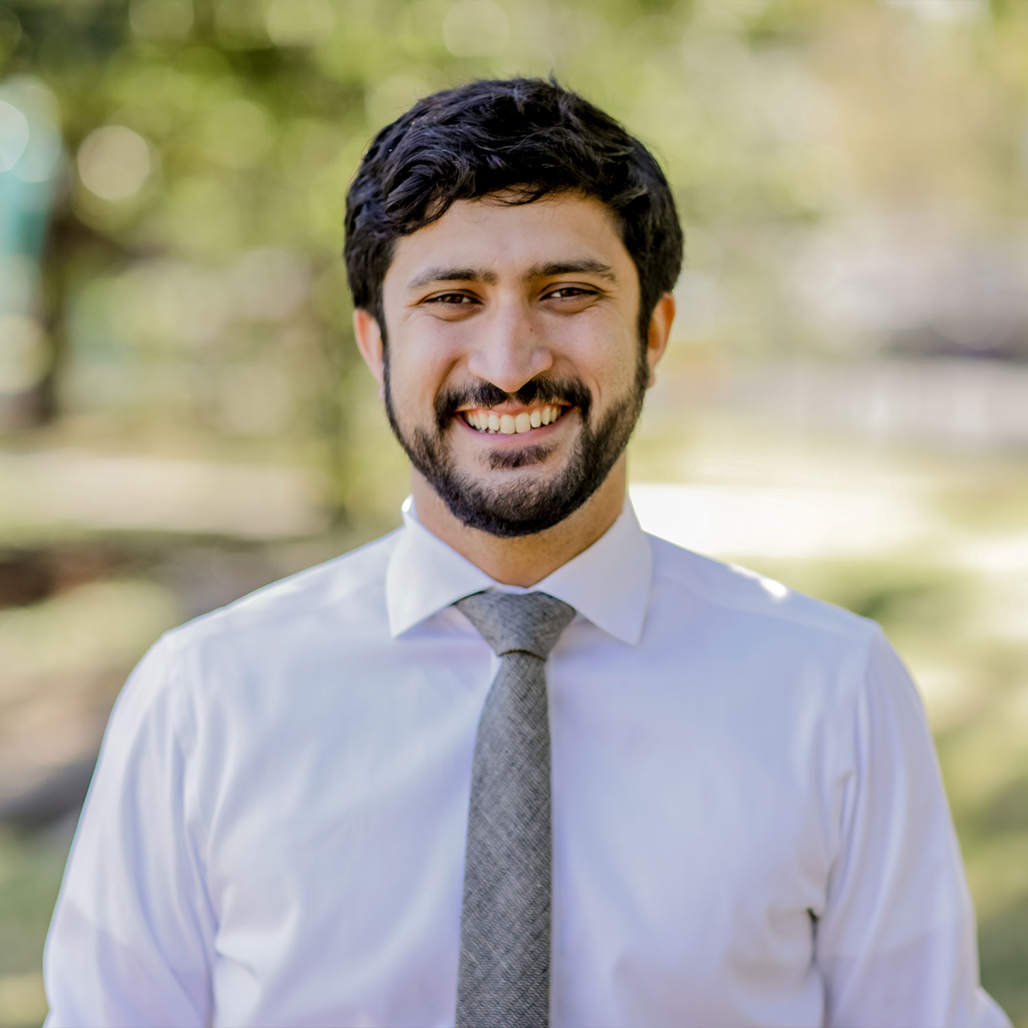 Greg Casar is shown smiling outdoors, wearing a white dress shirt and gray tie, with a blurred natural background.