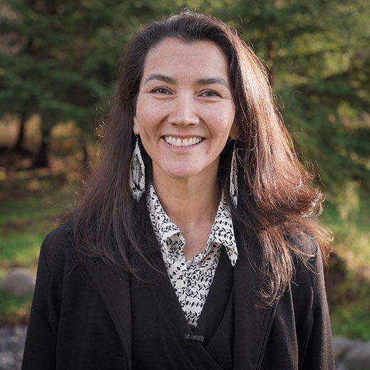 Mary Peltola smiling warmly outdoors, with long dark hair, wearing a dark coat over a patterned blouse, and standing in front of a peaceful natural background of green trees.