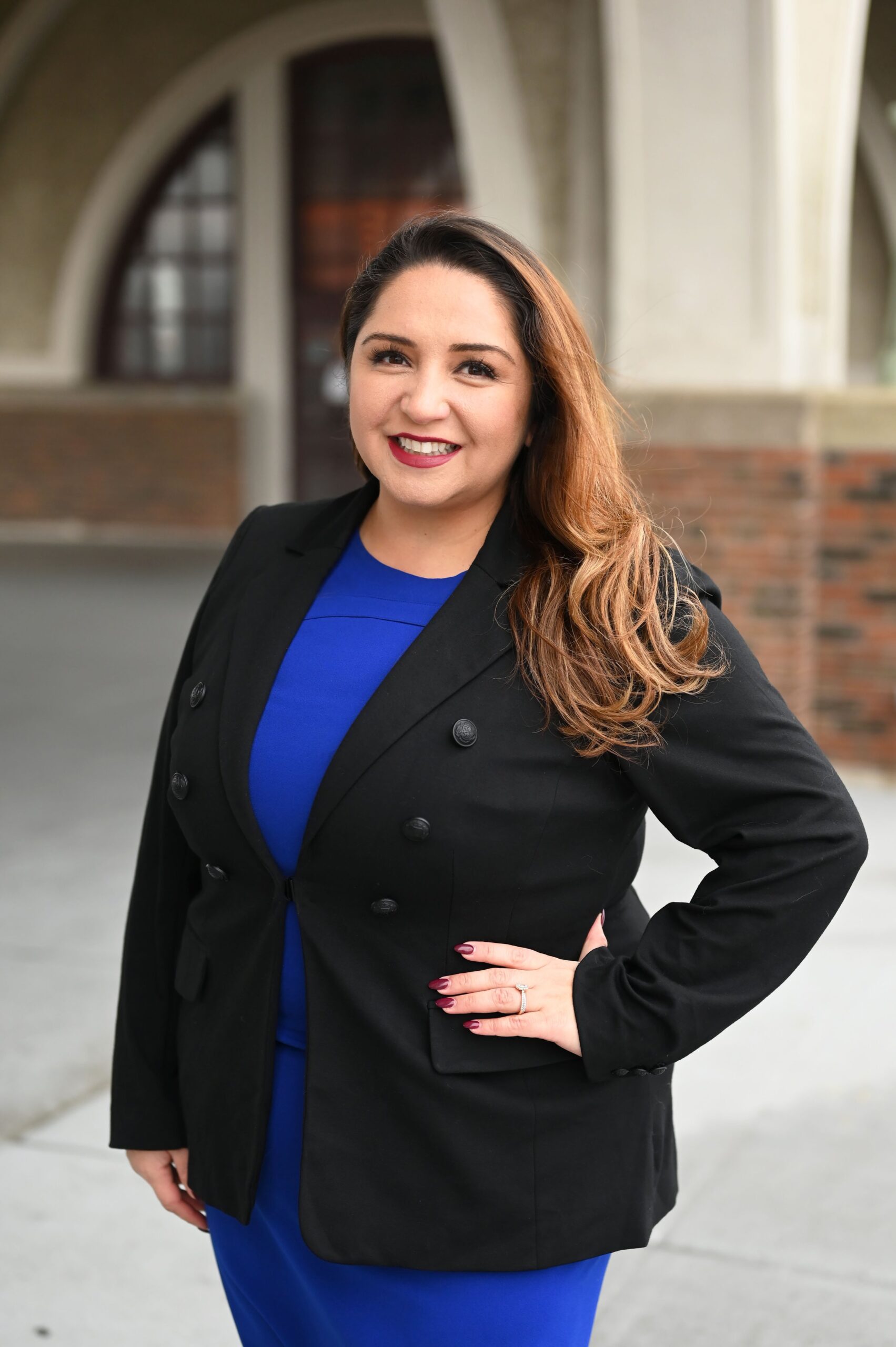 Delia Ramirez is pictured smiling confidently in this image, wearing a blue dress and a black blazer, standing with her hand on her hip in front of a building with arched windows.