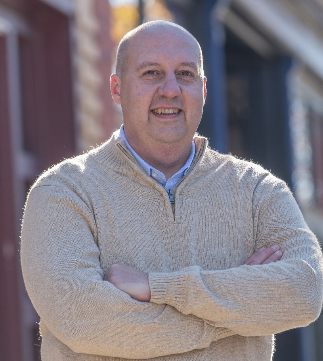 Curtis Hertel smiling confidently with arms crossed, wearing a beige sweater, and standing in front of a blurred street scene with buildings and autumn trees in the background.