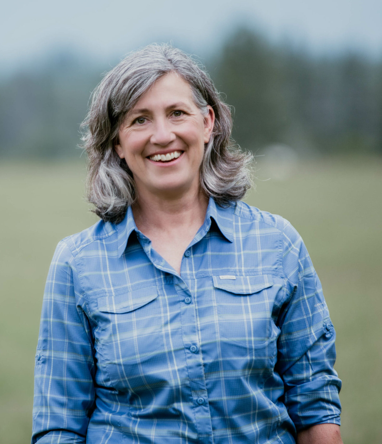 Monica Tranel is smiling outdoors in a blue plaid shirt against a blurred natural background, embodying her down-to-earth and approachable nature.