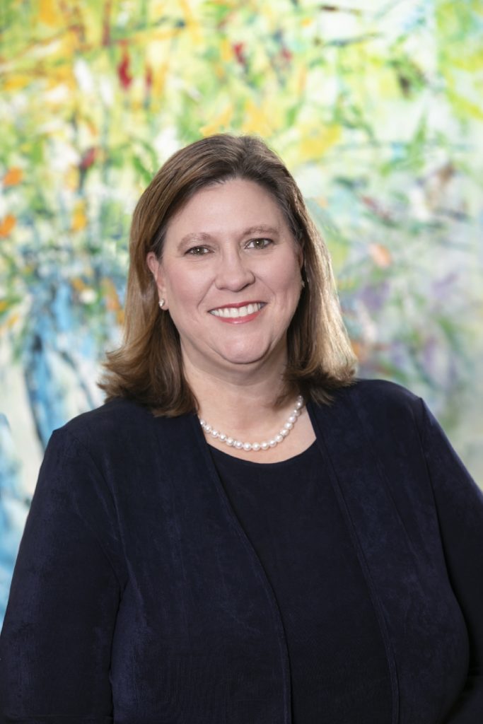 Julie Johnson is shown in this image wearing a dark outfit with a pearl necklace, smiling confidently in front of a vibrant, abstract background.