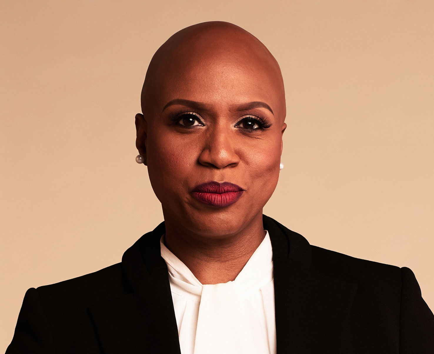 Ayanna Pressley is shown in this image wearing a black blazer and white blouse, with a confident expression, set against a neutral background.