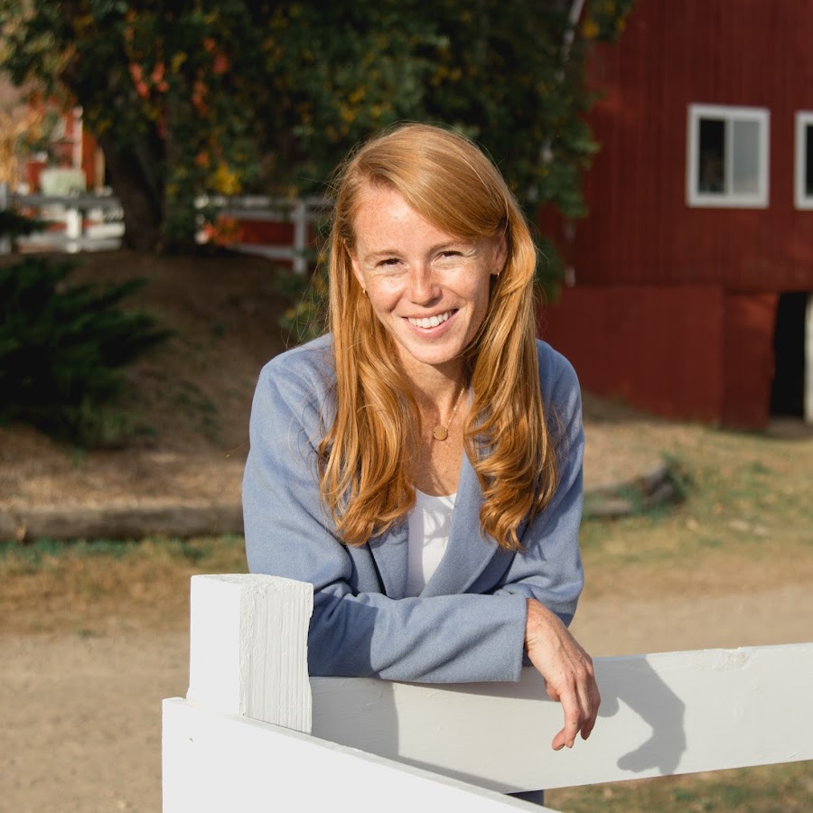 Rebecca Cooke smilling leaning over a fence with a barn-like background.