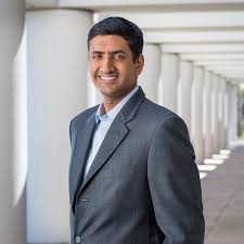 Ro Khanna smiling in a dark suit jacket, standing in front of a series of white columns in a shaded walkway, projecting a sense of professionalism and approachability.