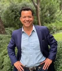 John Avlon, wearing a blue suit and light shirt, smiles confidently with his hands on his hips in a natural outdoor setting.