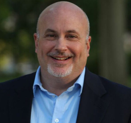 Mark Pocan is shown smiling outdoors in this image, wearing a light blue shirt and a dark blazer with a blurred green background.