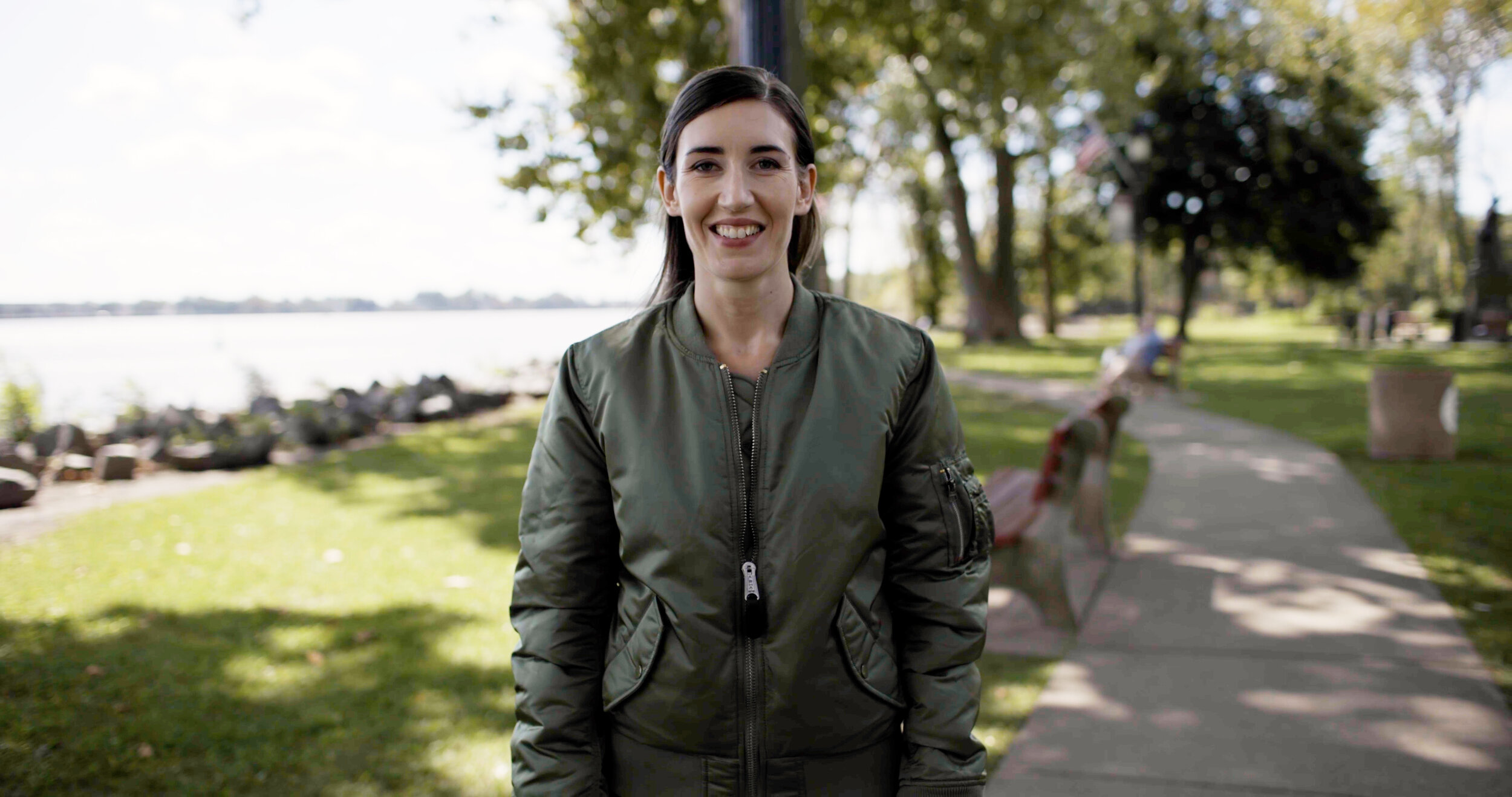 Ashley Ehasz is pictured outdoors in a green jacket, standing in a peaceful park setting with a serene water backdrop.