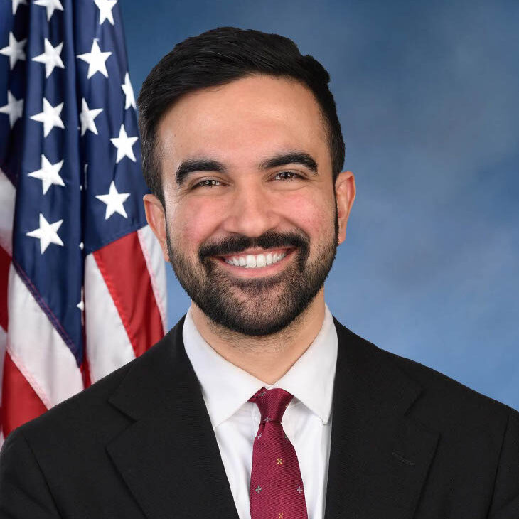 Zohran Mamdani for NYC Mayor portrait smiling at the camera wearing a black suit with red tie. He is posed with a blue background and an American flag behind his right shoulder.